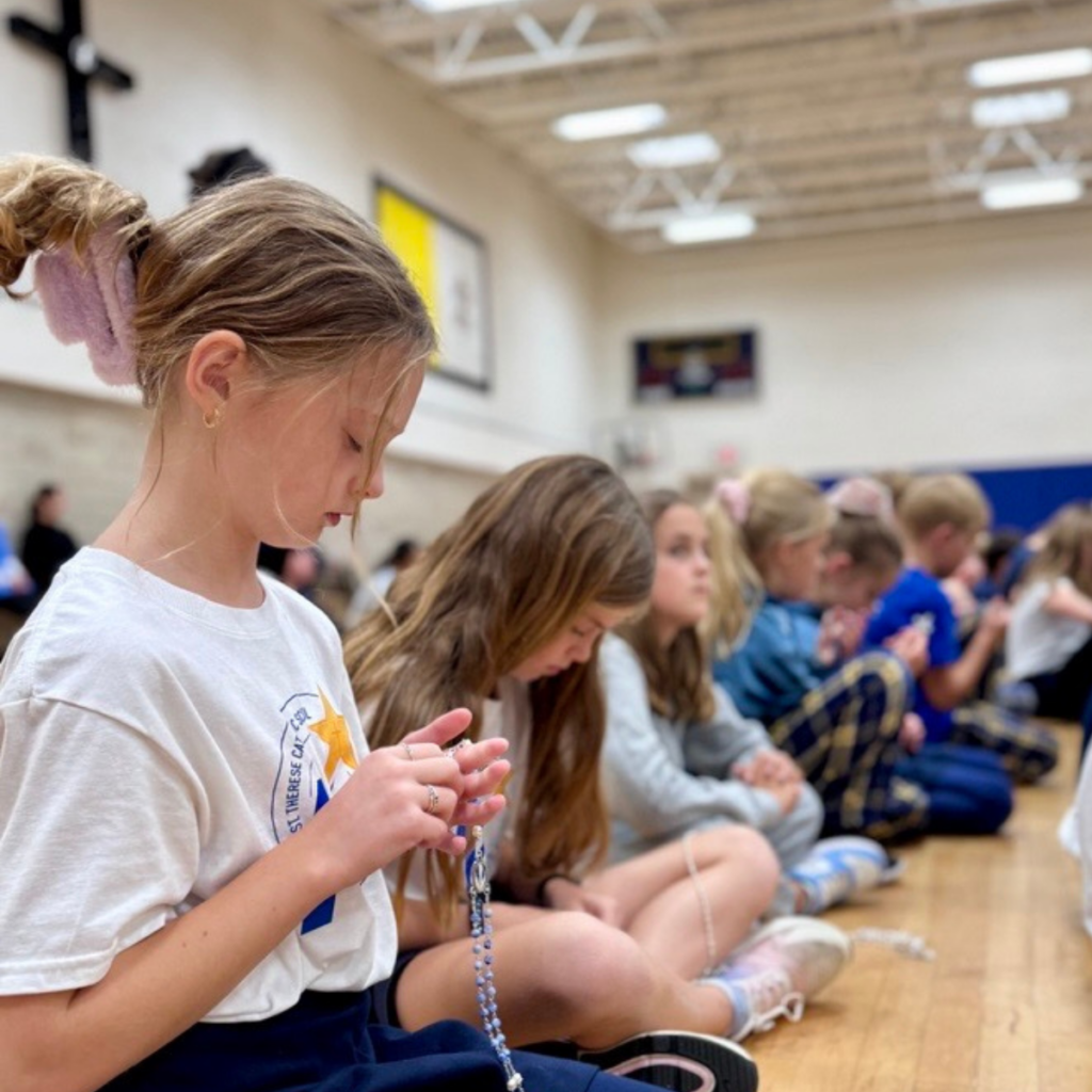 Children sitting in gymnasium holding beaded necklaces during school event.