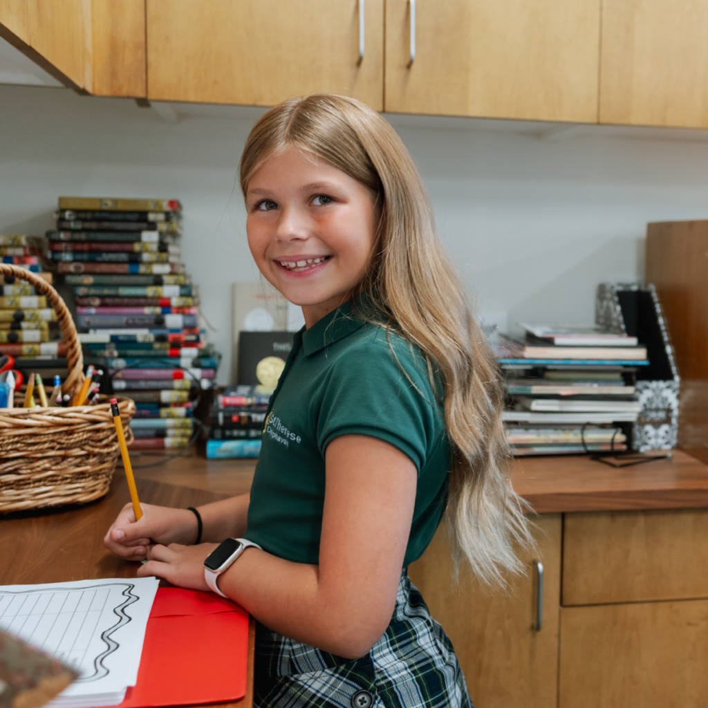 Smiling student at classroom desk with books and supplies behind her.