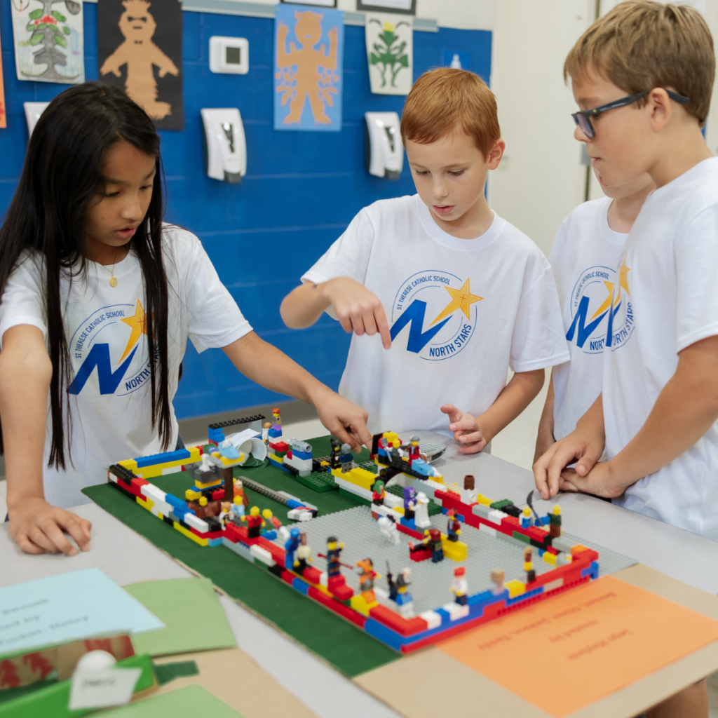 Three elementary students in matching school shirts building with colorful LEGO bricks at a table.